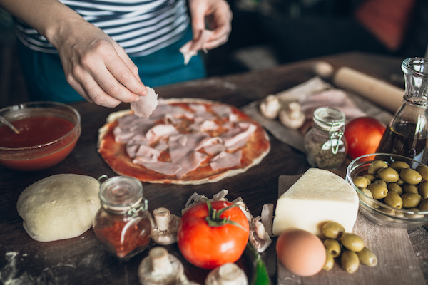 Young woman making pizza in domestic kitchen at home, with traditional pizza ingredients