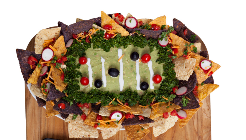 An overhead extreme close up horizontal photograph of a large wooden cutting board with some super bowl snacks consisting of a tray of guacamole and chips decorated to look like a football playing field and spicy buffalo wings on a grungy old baking tray