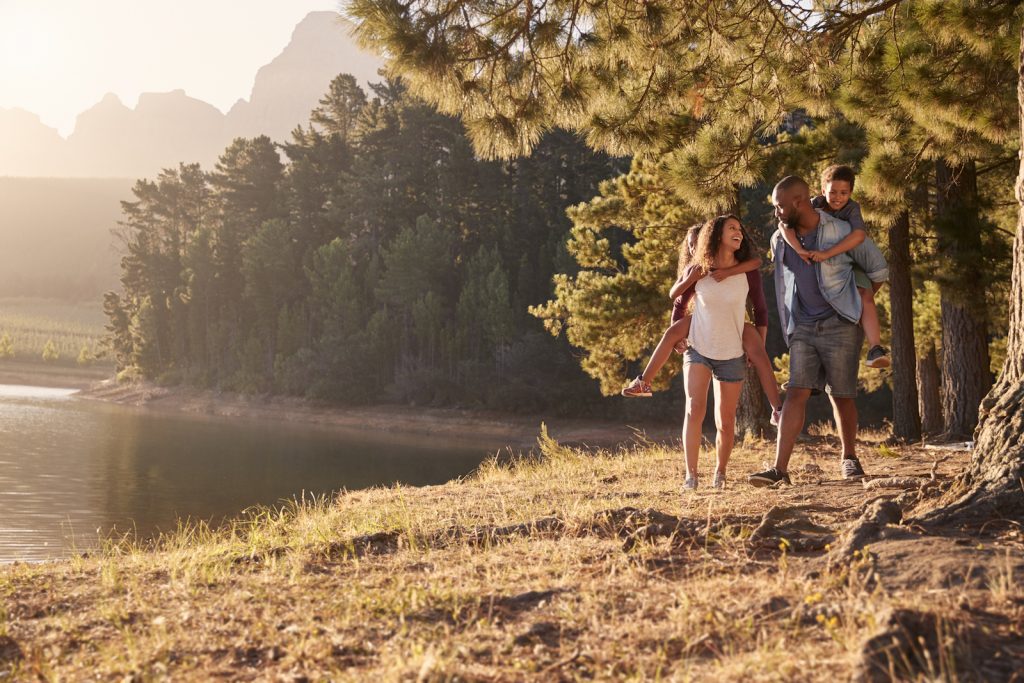 Parents Giving Children Piggyback Rides On Walk By Lake during Summer Vacation trip.