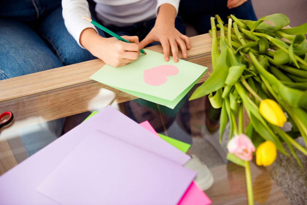 Girl's hands writing wish on handmade postcard near bouquet of yellow fresh tulips.