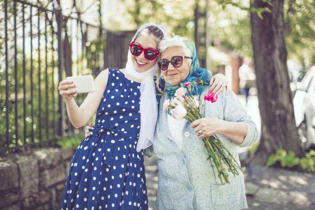 Smiling young teenager girl is taking selfies of herself and her grandmother while the woman holds a gift and a bunch of flowers.