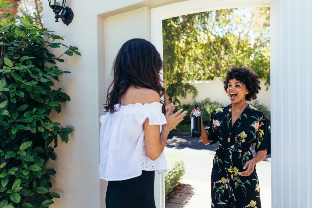 Excited friend attending a friend's housewarming party and giving a wine bottle to her friend at entrance door.