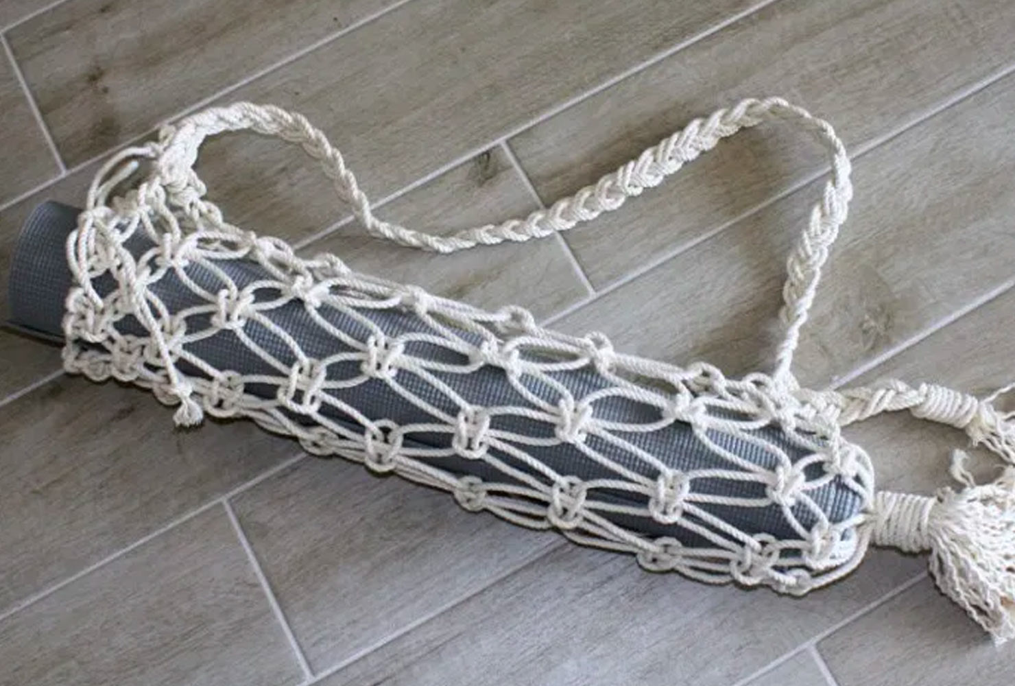 Macrame yoga mat carrier sitting on floor holding gray mat.